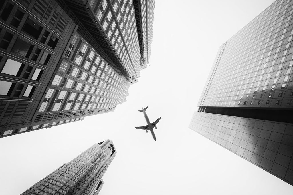 Schwarz-Weiß Fotografie von einem Flugzeug das über Hochhäuser fliegt mit Blick von unten. Fotokunst online kaufen. Wandbild hinter Acrylglas oder als Poster