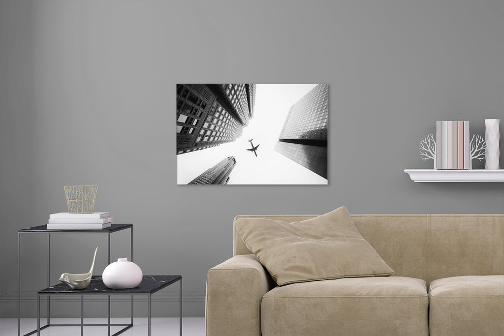 Aufgehängte Schwarz-Weiß Fotografie von einem Flugzeug das über Hochhäuser fliegt mit Blick von unten. Fotokunst online kaufen. Wandbild hinter Acrylglas oder als Poster