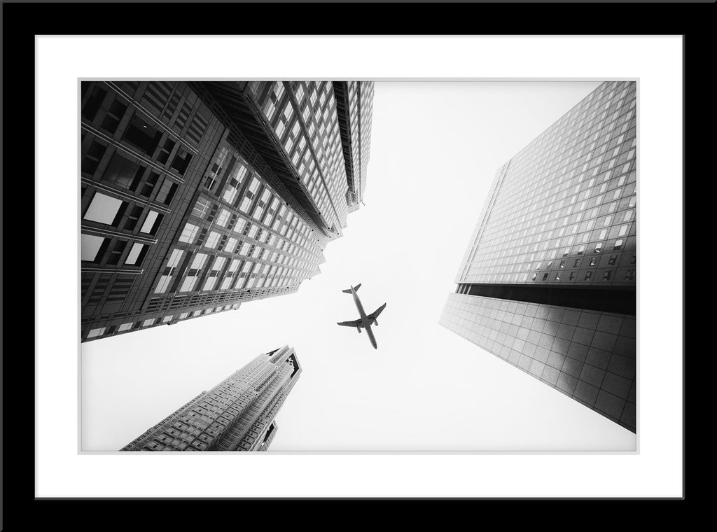 Schwarz-Weiß Fotografie von einem Flugzeug das über Hochhäuser fliegt mit Blick von unten. Fotokunst online kaufen. Wandbild im Rahmen