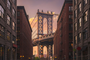 Stadt Architektur Fotografie der Manhattan Bridge durch eine Häuserfront bei Sonnenuntergang in New York. Fotokunst und Bilder online kaufen. Wandbild hinter Acrylglas oder als Poster