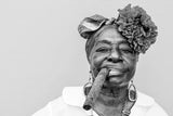 Schwarz-Weiß People Fotografie von einer dunkelhäutigen älteren Dame mit Blumen um Haar die eine decke Zigarre raucht. Fotokunst und Bilder online kaufen. Wandbild hinter Acrylglas oder als Poster