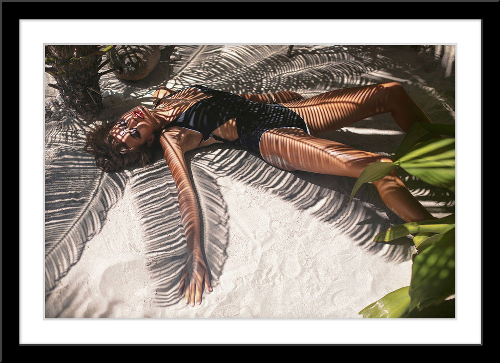 People Fotografie von einer Frau im schwarzen Badeanzug die am Stand im Schatten einer Palme liegt. Fotokunst und Bilder online kaufen. Wandbild im Rahmen